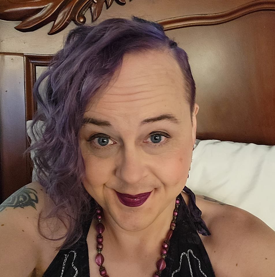 Zoe headshot - a woman with purple shoulder length hair swept to one side and a sideshave on the other, she is caucasian with blue eyes and a smirk and purple lipstick, sitting in front of an ornate headboard for a bed. 